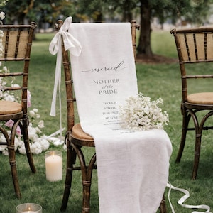 May include: A wedding chair with a white linen runner, bearing the text "Reserved Mother of the Bride." The chair is decorated with a small bouquet of white flowers and a white ribbon. Other chairs and candles are visible, set on a grassy area.