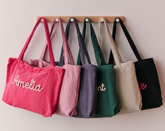 May include: A selection of corduroy tote bags in shades of pink, purple, green, and black, suspended from a wooden rail. Each bag has embroidered text, with the name "Amelia" visible on the pink bag.