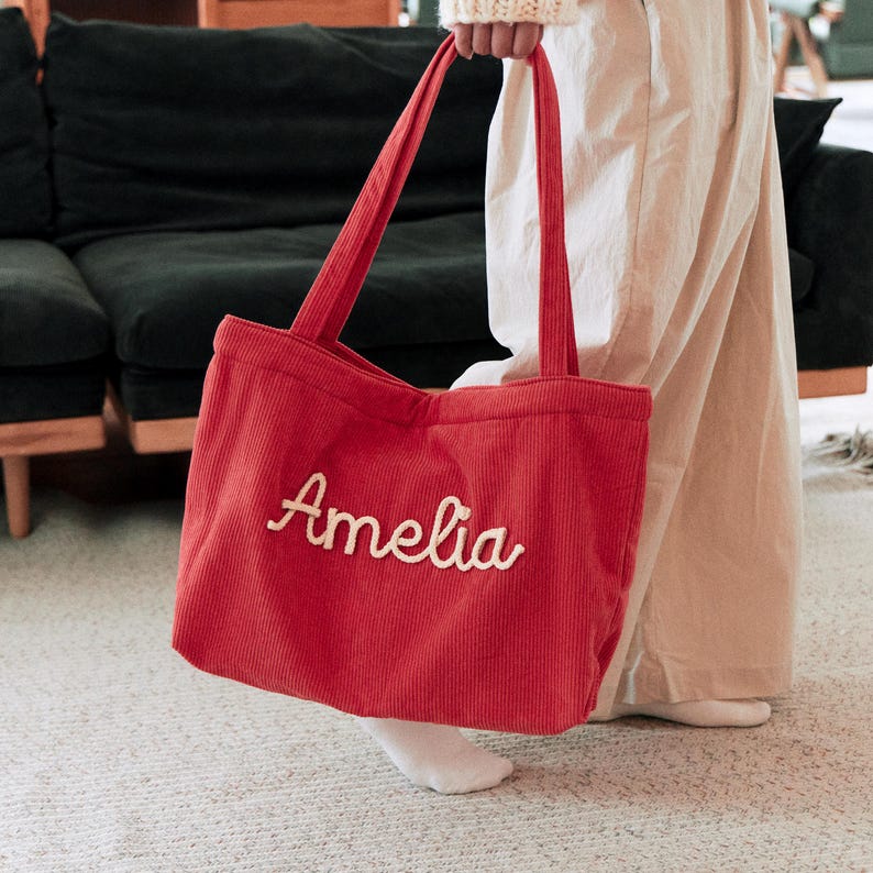 May include: A red corduroy tote bag featuring two long shoulder straps and the name Amelia embroidered in white cursive script across the front. The bag has a textured ribbed fabric surface and a rectangular shape with a spacious main compartment.