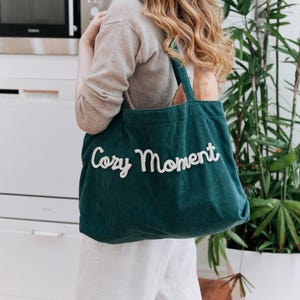 May include: A dark green corduroy tote bag featuring the text "Cozy Moment" embroidered in white cursive lettering across the front. The bag has two shoulder straps and a spacious rectangular body, providing a soft, textured appearance.