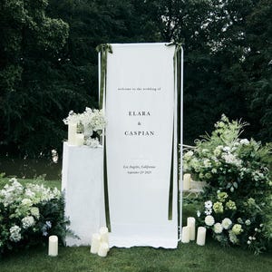 May include: A white wedding welcome sign with the text "welcome to the wedding of ELARA & CASPIAN" and the location and date. The sign is decorated with green ribbons and surrounded by white flowers and candles on a green lawn.