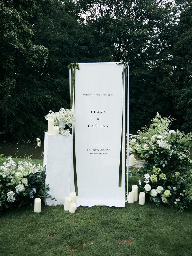 May include: A white wedding welcome sign with the text "welcome to the wedding of ELARA & CASPIAN" and the location and date. The sign is decorated with green ribbons and surrounded by white flowers and candles on a green lawn.