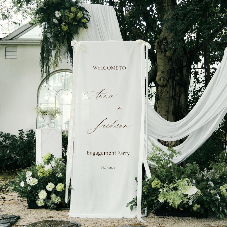 May include: A white fabric banner with the words "WELCOME TO" above the names "Anna" and "Jackson" in elegant script. The banner also reads "ENGAGEMENT PARTY" and the date "03.07.2025". The banner is tied with white ribbons and surrounded by greenery.