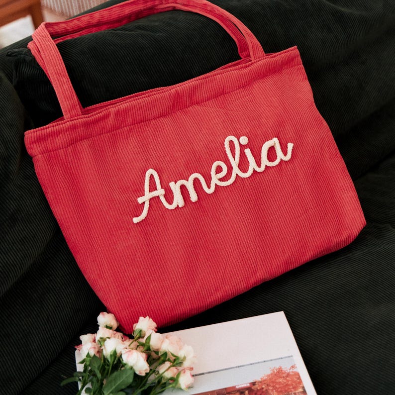 May include: A red corduroy tote bag featuring two shoulder straps and the name Amelia embroidered in white cursive script across the front centre. The fabric has a distinct vertical ribbed texture throughout the entire surface of the bag.