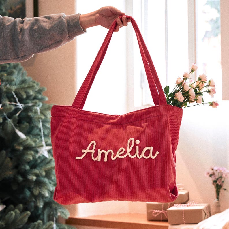 May include: A red corduroy tote bag featuring two long shoulder straps and the name Amelia embroidered in white cursive script on the front. The bag has a textured ribbed surface and is shown with small pink flowers tucked inside the opening.