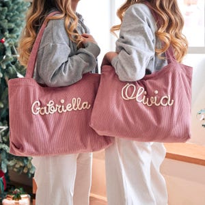 May include: Two dusty rose corduroy tote bags featuring embroidered names in a cream-coloured script font. One bag displays the name Gabriella, and the other displays the name Olivia. Each bag has two shoulder straps and a textured fabric surface.
