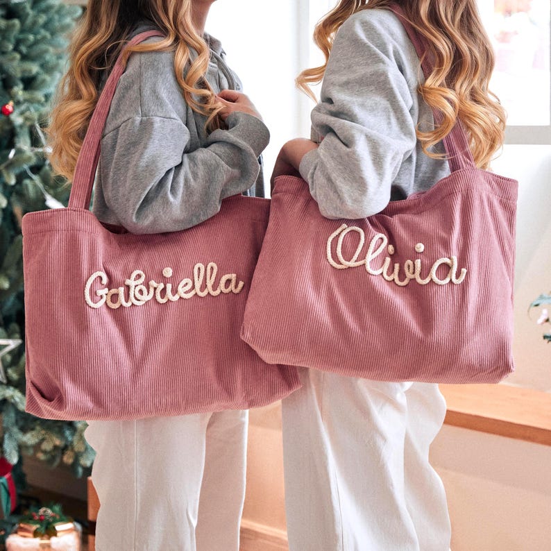 May include: Two dusty rose corduroy tote bags featuring embroidered names in a cream-coloured script font. One bag displays the name Gabriella, and the other displays the name Olivia. Each bag has two shoulder straps and a textured fabric surface.