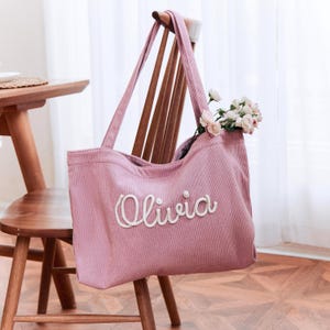 May include: A dusty pink corduroy tote bag featuring the name Olivia embroidered in white cursive script on the front. The bag has two shoulder straps and contains a small bouquet of light pink and white roses tucked inside the main opening.