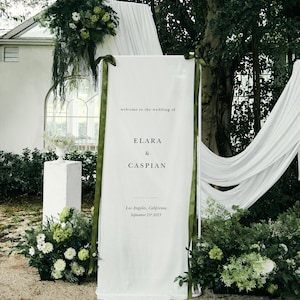 May include: A white wedding welcome sign with the names ELARA & CASPIAN, and the date September 21st, 2025. The sign is decorated with green ribbons and surrounded by greenery and white flowers, creating an elegant outdoor setting.
