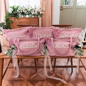 May include: Five dusty pink corduroy tote bags with two handles each, featuring white embroidered names Jessie, Margot, Gabriella, Natalie, and Olivia. Each bag is adorned with a small pink ribbon and white floral accents on the front.