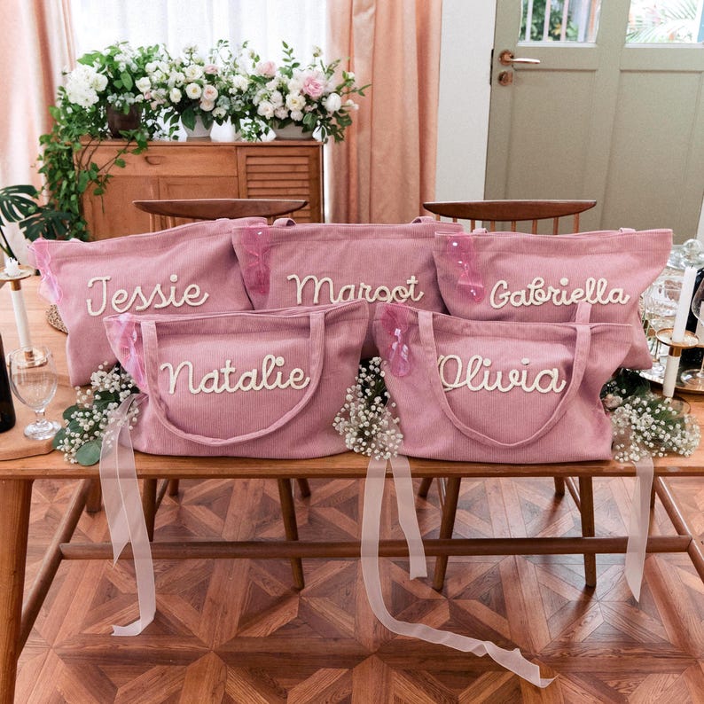 May include: Five dusty pink corduroy tote bags with two handles each, featuring white embroidered names Jessie, Margot, Gabriella, Natalie, and Olivia. Each bag is adorned with a small pink ribbon and white floral accents on the front.