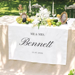 May include: A white table runner featuring the printed text MR & MRS. Bennett and the date 13.07.2026 in black font. The runner is draped over a beige tablecloth, decorated with a centerpiece of white flowers, green grapes, lemons, and limes.