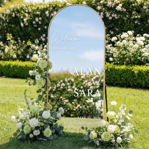 May include: A large, arched mirror with a gold frame, displaying the text "Welcome" and the names "ALAN and SARA" with the date "MAY 31, 2026". The mirror is decorated with floral arrangements of white and green roses, set on a grassy lawn with a hedge background.