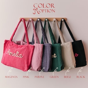 May include: Six corduroy tote bags hanging on a wooden rack, labelled COLOR OPTION. The bags are magenta, pink, purple, green, beige, and black. The magenta bag features the name Amelia in white script, while other bags show partial text.