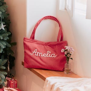 May include: A rose-coloured corduroy tote bag featuring two shoulder straps and the name Amelia embroidered in white cursive script across the front centre. The bag has a rectangular shape with a textured fabric surface and a top opening.
