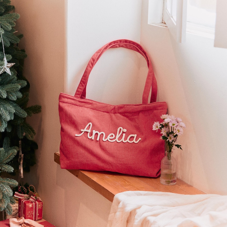 May include: A rose-coloured corduroy tote bag featuring two shoulder straps and the name Amelia embroidered in white cursive script across the front centre. The bag has a rectangular shape with a textured fabric surface and a top opening.
