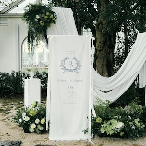 May include: A white banner with the text "LOVE IS THE HEAAOS" and the initials "H S" inside a floral wreath. The banner also includes the names "Hanna & Simon" and the date "06 23 25". The banner is part of a wedding ceremony setup.