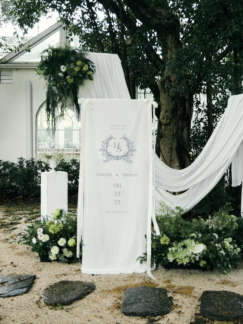 May include: A white banner with the text "LOVE IS THE HEAAOS" and the initials "H S" inside a floral wreath. The banner also includes the names "Hanna & Simon" and the date "06 23 25". The banner is part of a wedding ceremony setup.
