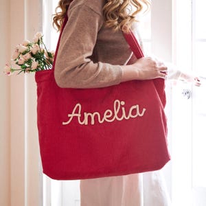 May include: A large red corduroy tote bag featuring the name Amelia embroidered in white cursive script on the front. The bag has two shoulder straps and contains a bouquet of light pink roses visible at the top opening of the main compartment.