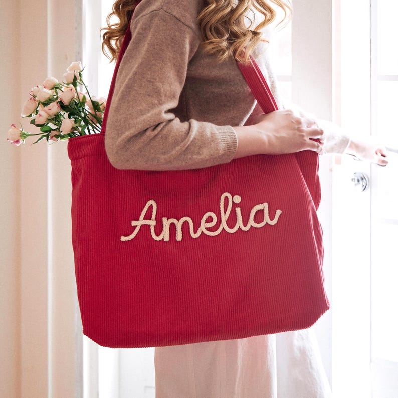 May include: A large red corduroy tote bag featuring the name Amelia embroidered in white cursive script on the front. The bag has two shoulder straps and contains a bouquet of light pink roses visible at the top opening of the main compartment.