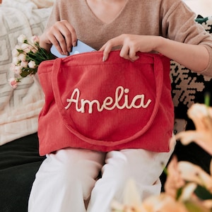 May include: A red corduroy tote bag featuring the name Amelia embroidered in white script lettering across the front. The bag has two matching red corduroy shoulder straps and a rectangular shape with a textured, ribbed fabric surface.