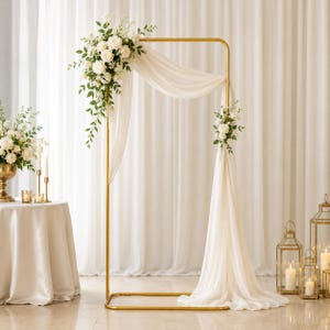 May include: A rectangular gold metal frame stand draped with sheer white fabric and decorated with white floral arrangements featuring roses and greenery. The structure is positioned upright, creating a decorative archway for event displays.