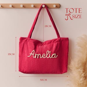 May include: A bright pink corduroy tote bag with the name Amelia embroidered in white script on the front. The bag features two shoulder straps and dimensions of 35 cm in height, 50 cm in width, and a 28 cm strap drop, labelled as TOTE SIZE.