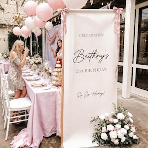 May include: A tall, rectangular, off-white fabric banner featuring the text CELEBRATING, Beithny's, 21st BIRTHDAY, and Sip Sip Hooray! in black script and sans-serif fonts. The banner is accented with dusty pink ribbons and white floral arrangements.