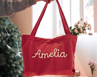 May include: A red corduroy tote bag featuring the name Amelia embroidered in white script lettering across the front. The bag has two long shoulder straps and contains a small bouquet of light pink roses tucked inside the main compartment.