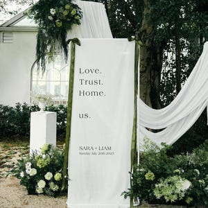 May include: A white wedding sign with the words "Love. Trust. Home. us." in black text. The sign is decorated with green ribbon and surrounded by greenery and flowers. The names "SARA + LIAM" and the date "Sunday July 20th 2025" are also displayed.