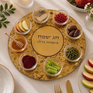 May include: A round gold-colored serving tray with a black floral pattern and Hebrew text that reads "Chag Sameach" and "Mishpachat Berlev". The tray holds six small glass bowls containing honey with a dipper, apple slices, fish, red berries, dates, green beans, and celery.