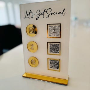 May include: White acrylic stand with gold trim and the text "Let's Get Social". The stand has four gold circles with icons and QR codes for social media.