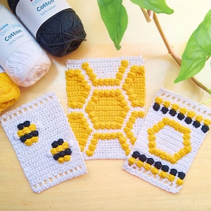 Può includere: Articoli all'uncinetto fatti a mano con un tema di api e favi. Gli articoli includono quadrati bianchi con disegni di api gialle e nere e un quadrato più grande con un motivo a nido d'ape giallo. Sono visibili anche gomitoli di lana bianchi, gialli e neri.