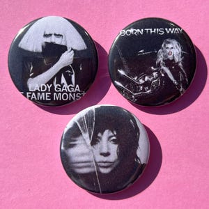 May include: Three black and white button badges featuring Lady Gaga. The first badge features Lady Gaga with her hair covering her face and the text "Lady Gaga Fame Monster". The second badge features Lady Gaga with the text "Born This Way". The third badge features Lady Gaga with her hair covering her face.