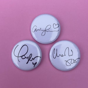 May include: Three white, circular badges featuring black handwritten signatures. The signatures are in cursive and include hearts and other small symbols. The badges are set against a pink backdrop.