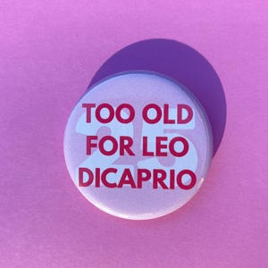May include: A circular pin-back button featuring a light pink background with the bold red text "TOO OLD FOR LEO DICAPRIO" and a large, faint white number "25" printed in the centre, creating a clear and readable graphic design on the surface.