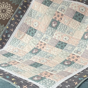 May include: A patchwork baby quilt in pastel shades. The quilt features squares with animal and number designs, including rabbits, bears, and the numbers 2 and 3. The border is grey with letters.