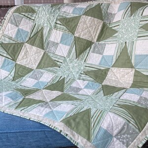 May include: A quilted blanket featuring a repeating star pattern in shades of sage green, light blue, and white. The design incorporates various fabric textures, including solid green triangles, striped panels, and small floral or dotted prints.