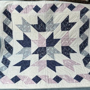 May include: A handmade quilt with a starburst design. The quilt features a white background with a central star pattern in navy blue and light pink. The border includes geometric shapes in coordinating colours. The quilt is likely made of cotton and is suitable for a bed or as a decorative throw.