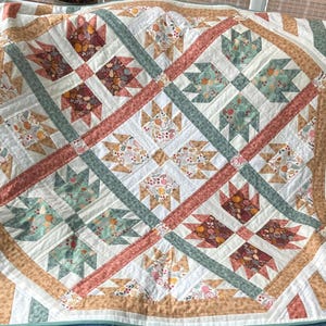 May include: A handmade quilt with a repeating pattern of bear paw blocks in shades of white, peach, sage green, and burgundy. The quilt features a diagonal grid design with a border of tan and white stripes. The overall theme is a cozy, rustic aesthetic.