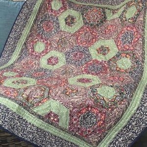 May include: A patterned quilt in pink, green, and blue. The quilt has a repeating hexagonal design with a green border and a dark blue floral edge. The quilt is folded over a blue cushion. The quilt's dimensions are approximately 150cm x 200cm.