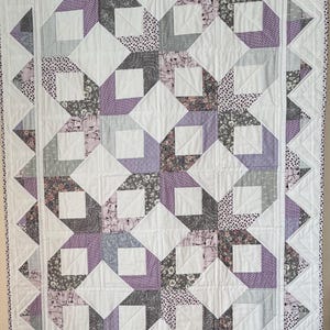 May include: A handmade quilt with a geometric pattern in white, lavender, grey, and floral fabrics. The quilt features square and diamond shapes, with a white border and a decorative edge.