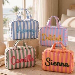 May include: Four rectangular toiletry bags with vertical stripes and top handles. The bags feature raised lettering in cursive script: blue bag with 'Avery', pink and blue bag with 'Delilah', green and pink bag with 'Camille', and orange and pink bag with 'Sienna'.