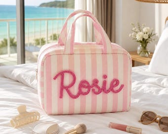 May include: A rectangular travel bag featuring vertical light pink and white stripes with the name Rosie embroidered in raised pink script on the front. The bag has two top handles and a zipper closure, accompanied by a hair clip, makeup brush, lipstick, and sunglasses.