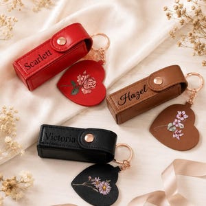 May include: Three rectangular lipstick cases with snap closures and attached heart-shaped keychains. The red case is engraved with Scarlett and a rose, the brown case with Hazel and flowers, and the black case with Victoria and daisies.