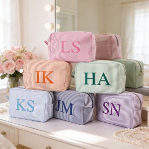 Puede incluir: Una colección de neceseres a rayas en varios colores, incluyendo rosa, naranja, azul y verde. Cada bolsa presenta una inicial bordada en un color contrastante. Las bolsas están dispuestas sobre una superficie blanca.