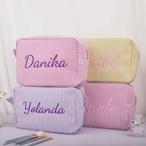 Peut inclure: Quatre trousses de maquillage personnalisées avec des motifs à rayures verticales roses, jaunes et violettes. Chaque sac porte un nom différent brodé dans une couleur complémentaire : Danika, Gabi, Yolanda et Jaelle. Les sacs sont rectangulaires avec une fermeture éclair.