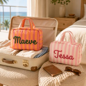 May include: Two rectangular travel pouches with top handles and vertical stripes. One pouch is orange and pink with the name Maeve in green script, and the other is white and pink with the name Tessa in pink script. Both feature zippered closures.