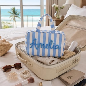 May include: A rectangular toiletry bag with light blue and white vertical stripes, featuring the name Amelia in blue cursive script on the front. The bag has two top handles and is placed inside an open beige suitcase with other travel items.
