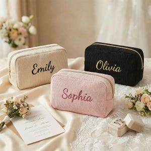 Puede incluir: Tres neceseres de pana en crema, rosa y negro, cada uno bordado con un nombre: Emily, Sophia y Olivia. Las bolsas tienen forma rectangular con cierre de cremallera. El fondo incluye flores y una invitación de boda.
